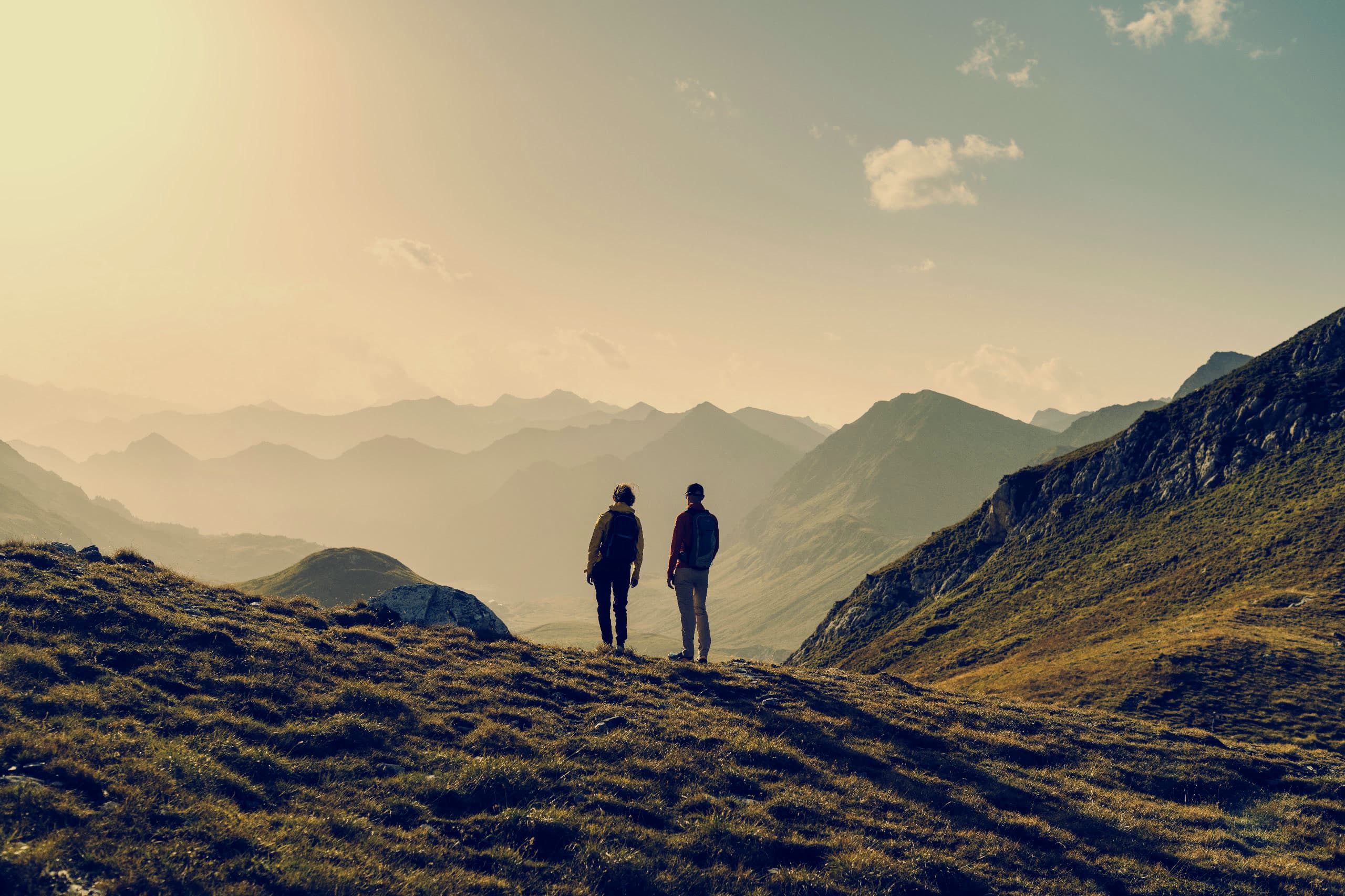 Two hikers on an alpine ridge at golden hour, looking out over layered Swiss mountains